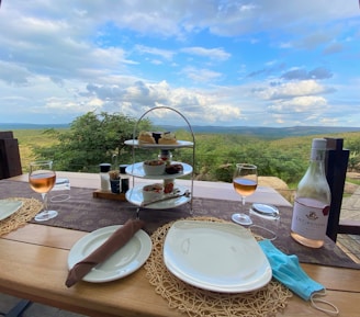 A picturesque outdoor dining setup features a table with two plates, a bottle of rosé, two glasses of wine, and a tiered serving stand with various appetizers and desserts. The table overlooks a lush, expansive landscape with rolling hills under a partly cloudy sky.