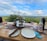 A picturesque outdoor dining setup features a table with two plates, a bottle of rosé, two glasses of wine, and a tiered serving stand with various appetizers and desserts. The table overlooks a lush, expansive landscape with rolling hills under a partly cloudy sky.