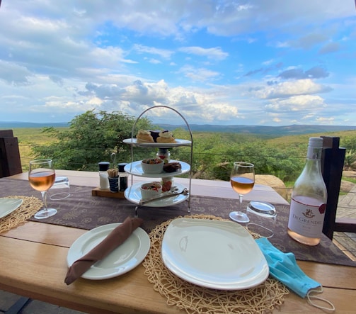 A picturesque outdoor dining setup features a table with two plates, a bottle of rosé, two glasses of wine, and a tiered serving stand with various appetizers and desserts. The table overlooks a lush, expansive landscape with rolling hills under a partly cloudy sky.