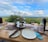 A picturesque outdoor dining setup features a table with two plates, a bottle of rosé, two glasses of wine, and a tiered serving stand with various appetizers and desserts. The table overlooks a lush, expansive landscape with rolling hills under a partly cloudy sky.