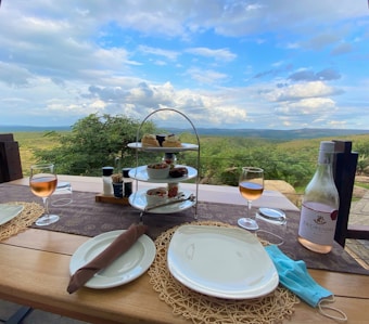 A picturesque outdoor dining setup features a table with two plates, a bottle of rosé, two glasses of wine, and a tiered serving stand with various appetizers and desserts. The table overlooks a lush, expansive landscape with rolling hills under a partly cloudy sky.