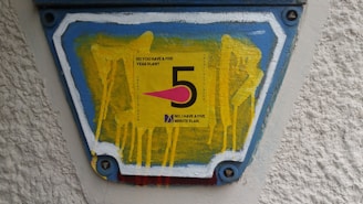 A colorful sign featuring the number '5' prominently in black on a yellow background. Surrounding the number is a question, 'Do you have a five year plan?' and beneath it, a statement, 'No, I have a five minute plan.' The sign is mounted on a textured surface with blue, white, and red borders containing yellow drips of paint.