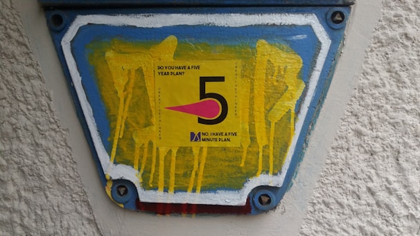 A colorful sign featuring the number '5' prominently in black on a yellow background. Surrounding the number is a question, 'Do you have a five year plan?' and beneath it, a statement, 'No, I have a five minute plan.' The sign is mounted on a textured surface with blue, white, and red borders containing yellow drips of paint.