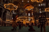 A grand, ornate interior with large, hanging chandeliers and high, domed ceilings. Arabic calligraphy adorns the walls, and numerous people are observing or exploring the space. The warm glow of the lighting creates a historical and majestic atmosphere.