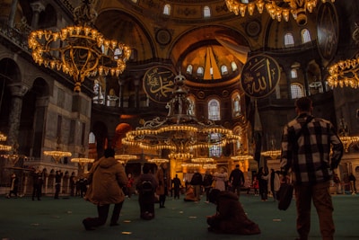 A grand, ornate interior with large, hanging chandeliers and high, domed ceilings. Arabic calligraphy adorns the walls, and numerous people are observing or exploring the space. The warm glow of the lighting creates a historical and majestic atmosphere.