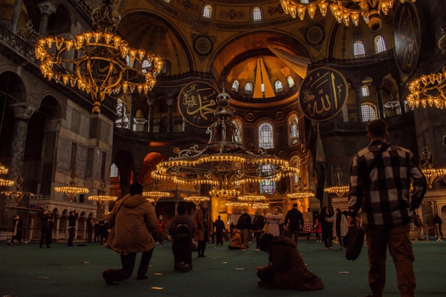 A grand, ornate interior with large, hanging chandeliers and high, domed ceilings. Arabic calligraphy adorns the walls, and numerous people are observing or exploring the space. The warm glow of the lighting creates a historical and majestic atmosphere.