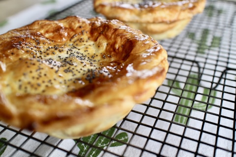 Golden-brown pastries topped with poppy seeds rest on a cooling rack. The background features a patterned cloth beneath the rack.