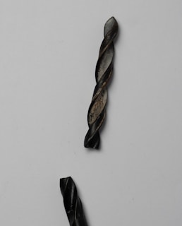 A close-up view of two black metallic twisted rods set against a plain light background. The rods have a spiral or drill bit-like pattern and show some wear and texture on their surfaces, suggesting use or oxidation.