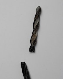 A close-up view of two black metallic twisted rods set against a plain light background. The rods have a spiral or drill bit-like pattern and show some wear and texture on their surfaces, suggesting use or oxidation.