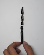 A broken drill bit is placed against a white background. The upper part of the bit aligns with the lower part, which is held by a hand with visible fingers.