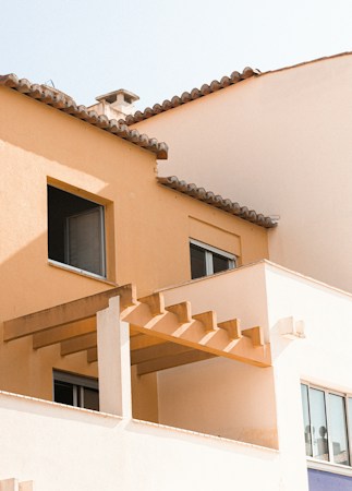 An architectural structure with a sunny, Mediterranean style features a light peachy-orange stucco facade and terracotta roof tiles. Large rectangular windows provide a modern touch, and a simple pergola adds a decorative element to the balcony.