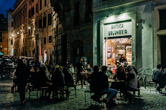 A charming evening scene depicts a bustling outdoor cafe setting in front of an old delicatessen named 'Antica Salumeria.' The street is lined with historical buildings under dim, warm street lighting. Groups of people are seen enjoying their meals and conversations at tables on the cobbled sidewalk. Inside the shop, shelves are filled with various goods, and a shopkeeper stands behind the counter.