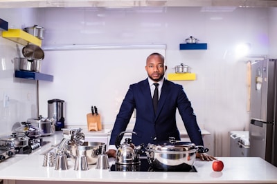 A man in a formal suit stands confidently in a modern kitchen. The kitchen is equipped with shiny stainless steel utensils and cookware. Shelves have colorful accents, with a mix of blue and yellow, complementing the overall sleek design. An apple is placed on the countertop near the man.