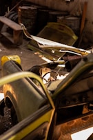 A dismantled and abandoned vehicle is situated in a dimly lit garage. The vehicle's hood is removed, exposing its internal components. Rusty barrels and a bench are in the background, along with other discarded items. Sunlight filters in, casting shadows across the dusty floor.