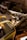 A dismantled and abandoned vehicle is situated in a dimly lit garage. The vehicle's hood is removed, exposing its internal components. Rusty barrels and a bench are in the background, along with other discarded items. Sunlight filters in, casting shadows across the dusty floor.