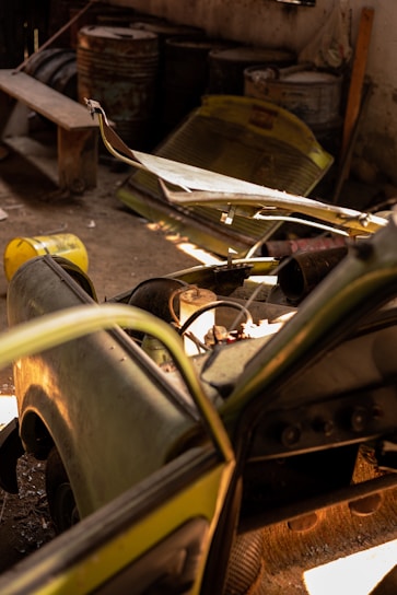 A dismantled and abandoned vehicle is situated in a dimly lit garage. The vehicle's hood is removed, exposing its internal components. Rusty barrels and a bench are in the background, along with other discarded items. Sunlight filters in, casting shadows across the dusty floor.