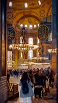 A bustling interior scene with a large group of people gathered inside a grand historical building. The space is adorned with intricate chandeliers, large decorative Arabic calligraphy on the walls, and ornate architectural details. The atmosphere is vibrant with activity as visitors explore the surroundings.