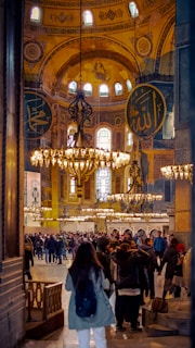 A bustling interior scene with a large group of people gathered inside a grand historical building. The space is adorned with intricate chandeliers, large decorative Arabic calligraphy on the walls, and ornate architectural details. The atmosphere is vibrant with activity as visitors explore the surroundings.