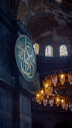 A large circular plaque with Arabic calligraphy is set against the interior of an ornate, historic building with arched windows. A grand chandelier with glowing lights hangs prominently in the foreground, casting a warm glow.