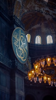 A large circular plaque with Arabic calligraphy is set against the interior of an ornate, historic building with arched windows. A grand chandelier with glowing lights hangs prominently in the foreground, casting a warm glow.