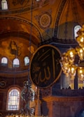 An intricately decorated interior with ornate Islamic calligraphy on a large circular panel. The architecture features high ceilings and detailed mosaics with religious symbols. Warm glowing lights hang from above, creating a serene and majestic atmosphere.