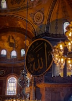 An intricately decorated interior with ornate Islamic calligraphy on a large circular panel. The architecture features high ceilings and detailed mosaics with religious symbols. Warm glowing lights hang from above, creating a serene and majestic atmosphere.