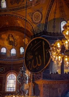 An intricately decorated interior with ornate Islamic calligraphy on a large circular panel. The architecture features high ceilings and detailed mosaics with religious symbols. Warm glowing lights hang from above, creating a serene and majestic atmosphere.