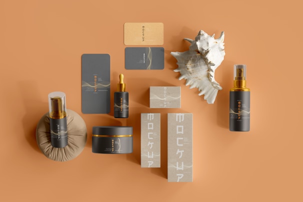 A collection of skincare products displayed on a tan background. The set includes various bottles and boxes featuring dark gray and beige colors with gold accents. A large seashell is placed among the products, adding an organic touch to the arrangement. The items are arranged neatly, highlighting their elegant design and branding.