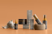Several skin care or cosmetics containers are arranged on a smooth orange surface. There are bottles with gold-accented caps, a jar, a small box-like structure, and a decorative seashell. The design is elegant and minimalist, with a consistent color scheme.