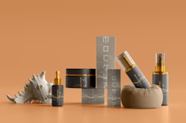 Several skin care or cosmetics containers are arranged on a smooth orange surface. There are bottles with gold-accented caps, a jar, a small box-like structure, and a decorative seashell. The design is elegant and minimalist, with a consistent color scheme.