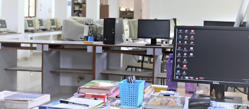 The image depicts an office or computer lab setting featuring a work desk in the foreground adorned with various stationery items, books, and a computer monitor displaying a desktop screen. In the background, there are multiple computer stations arranged in rows with older model monitors and workstations, suggesting a shared or communal workspace.