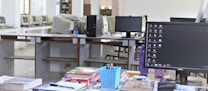 The image depicts an office or computer lab setting featuring a work desk in the foreground adorned with various stationery items, books, and a computer monitor displaying a desktop screen. In the background, there are multiple computer stations arranged in rows with older model monitors and workstations, suggesting a shared or communal workspace.