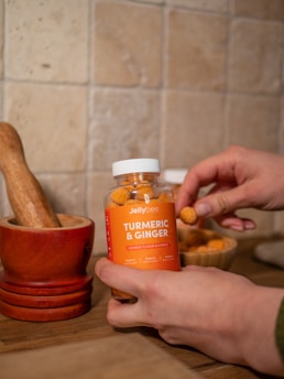 A person is holding a jar of orange flavored gummies labeled Turmeric & Ginger. The background includes a tiled wall and a wooden mortar and pestle set, creating a rustic kitchen ambiance. The jar is partially transparent, showing the gummies inside, and there is an additional bowl filled with gummies nearby.