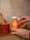 A person is holding a jar of orange flavored gummies labeled Turmeric & Ginger. The background includes a tiled wall and a wooden mortar and pestle set, creating a rustic kitchen ambiance. The jar is partially transparent, showing the gummies inside, and there is an additional bowl filled with gummies nearby.