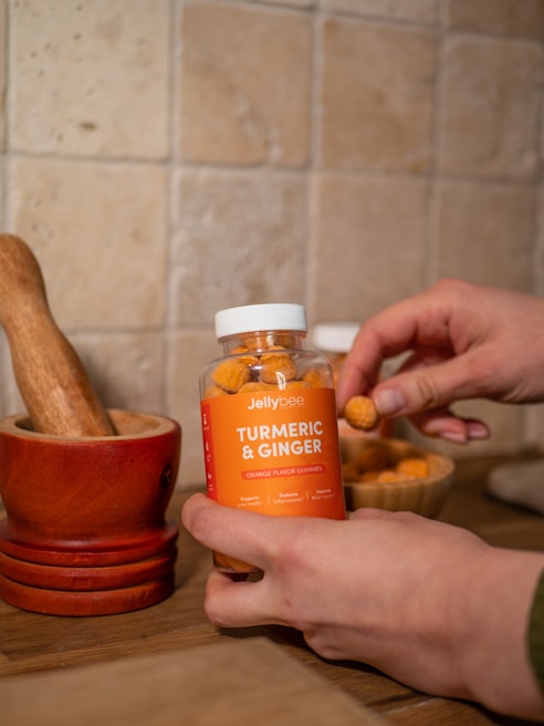A person is holding a jar of orange flavored gummies labeled Turmeric & Ginger. The background includes a tiled wall and a wooden mortar and pestle set, creating a rustic kitchen ambiance. The jar is partially transparent, showing the gummies inside, and there is an additional bowl filled with gummies nearby.