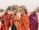 Four people are wearing traditional Maasai clothing, characterized by bright red and blue checked patterns. They stand close together, with arms linked, suggesting a sense of camaraderie. The background features a savanna landscape with sparse trees and dry grass.