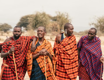Four people are wearing traditional Maasai clothing, characterized by bright red and blue checked patterns. They stand close together, with arms linked, suggesting a sense of camaraderie. The background features a savanna landscape with sparse trees and dry grass.