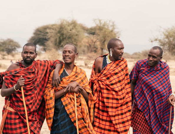 Four people are wearing traditional Maasai clothing, characterized by bright red and blue checked patterns. They stand close together, with arms linked, suggesting a sense of camaraderie. The background features a savanna landscape with sparse trees and dry grass.
