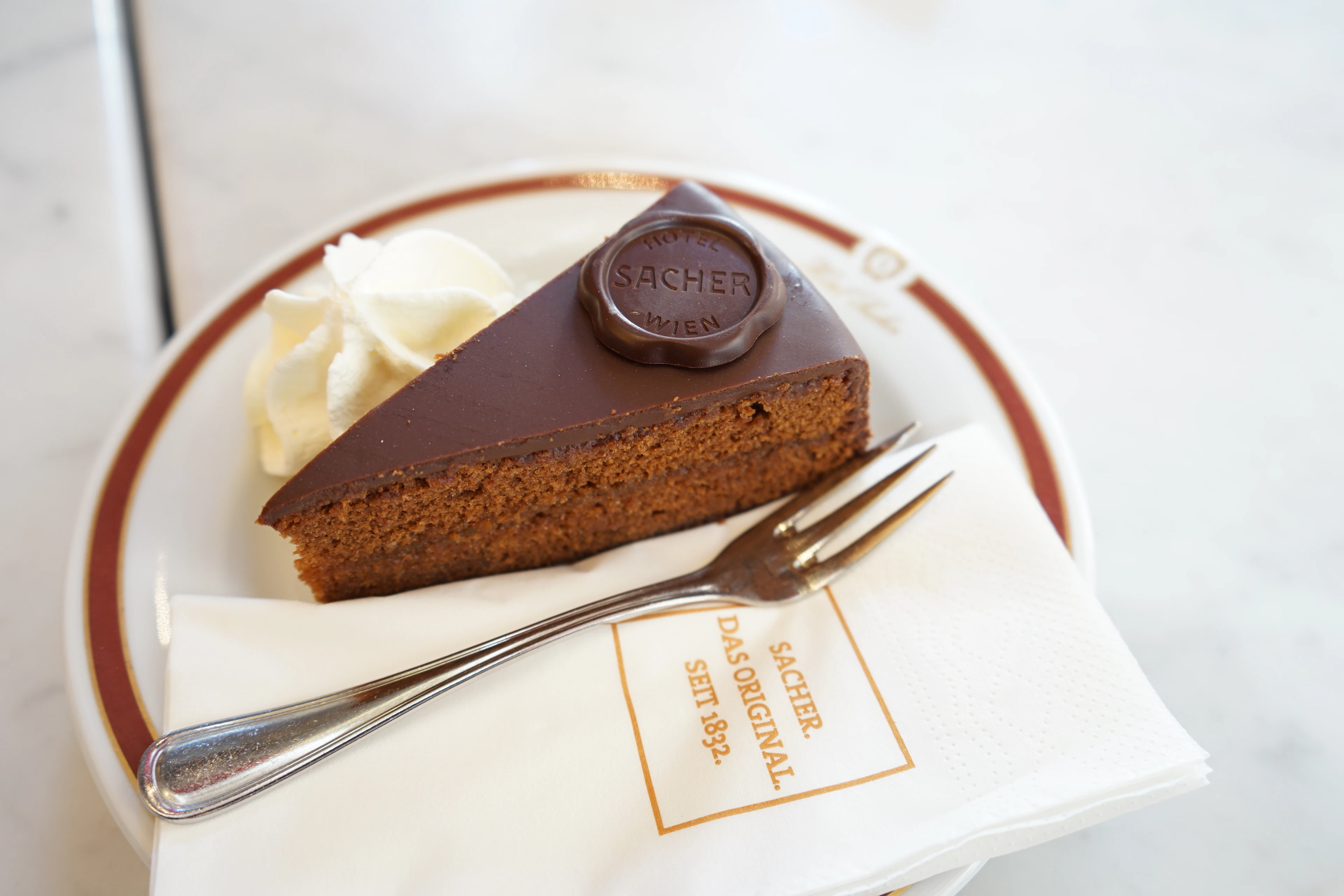 A slice of Original Sacher-Torte on a rimmed plate with a quenelle of unsweetened whipped cream, the round dark-chocolate seal stamped 'Sacher' on its glazed top, resting on a napkin printed with the Hotel Sacher mark.