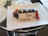A rectangular chocolate dessert topped with small pieces of gold leaf is placed on a white rectangular plate. The plate includes garnishes of sliced figs and blackberries. A white chocolate plaque with the words 'Happy Anniversary' written in chocolate is also placed on the dessert.