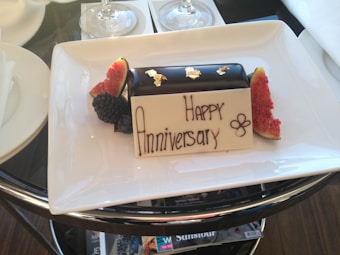 A rectangular chocolate dessert topped with small pieces of gold leaf is placed on a white rectangular plate. The plate includes garnishes of sliced figs and blackberries. A white chocolate plaque with the words 'Happy Anniversary' written in chocolate is also placed on the dessert.