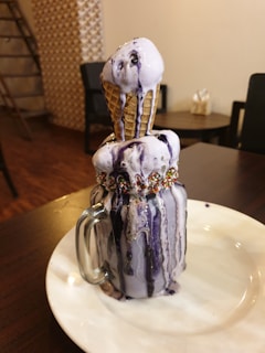 A decadent dessert featuring purple ice cream in a waffle cone placed upside down on top of a glass filled with more ice cream of the same flavor. The ice cream has melted slightly, with drips running down the sides. The glass is adorned with colorful sprinkles on the rim. It sits on a white plate in a cozy, dimly lit café setting.