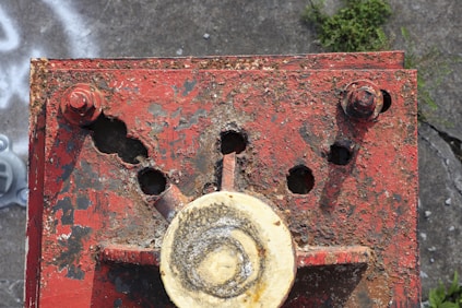 A corroded metal surface with several rusted bolts and circular holes, showing extensive wear and weathering. The metal is primarily red, with patches of rust and peeling paint, situated on a rough concrete surface.
