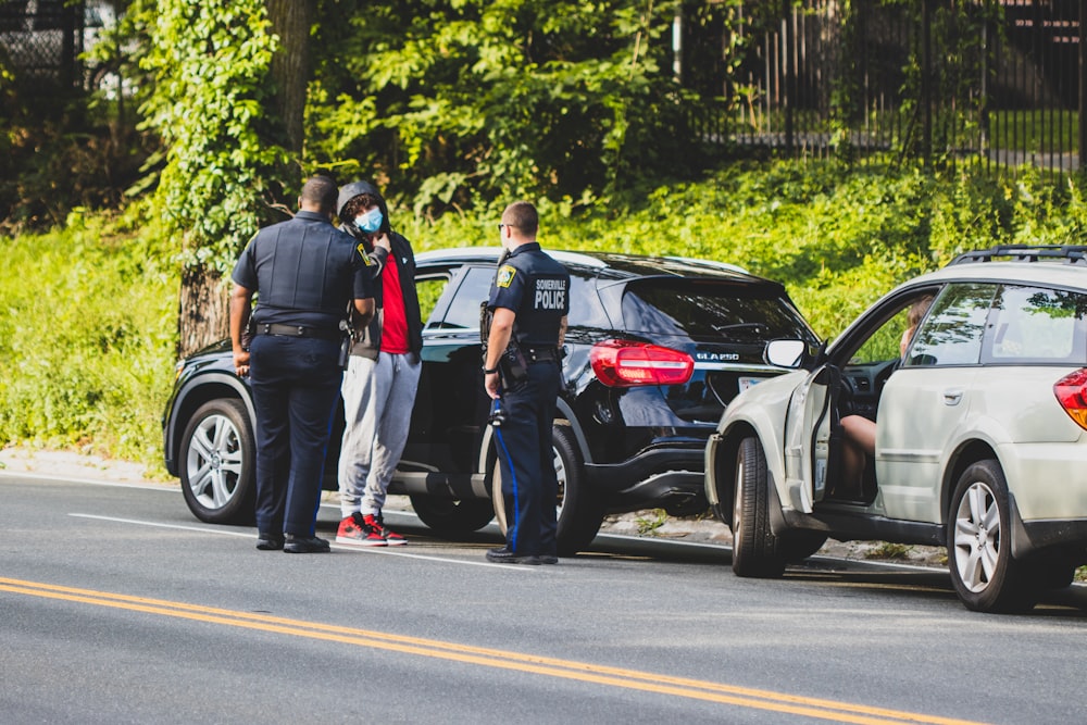 Hombre con camiseta negra y pantalones negros de pie junto a un SUV negro durante el día