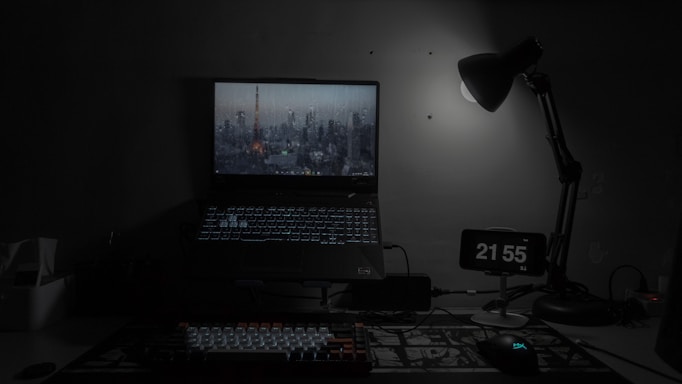 A dimly lit workspace features a laptop displaying a cityscape wallpaper, accompanied by a glowing keyboard and a computer mouse. An adjustable desk lamp casts light on a digital clock showing '21:55' with minimal decor visible around the setup.