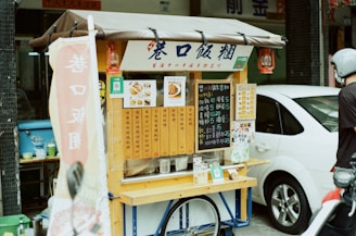 A small street food cart features a variety of menu options displayed on wooden panels and chalkboards. The cart is positioned on a street with a parked white car beside it. A person wearing a helmet is standing next to the cart. Red lanterns hang from the cart's awning, and a sign with Chinese characters hangs from the front.