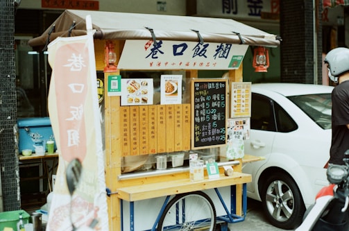 A small street food cart features a variety of menu options displayed on wooden panels and chalkboards. The cart is positioned on a street with a parked white car beside it. A person wearing a helmet is standing next to the cart. Red lanterns hang from the cart's awning, and a sign with Chinese characters hangs from the front.