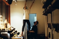 Two people are inside a room with one person standing on top of a counter or table reaching up to the ceiling, possibly performing maintenance or installation work. The other person is standing next to them, appearing to assist or observe. Both individuals are wearing masks. The room has shelves on the right side filled with various items and a counter or workspace area on the left.