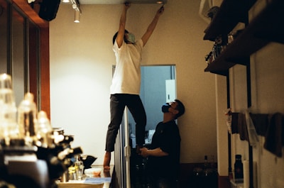 Two people are inside a room with one person standing on top of a counter or table reaching up to the ceiling, possibly performing maintenance or installation work. The other person is standing next to them, appearing to assist or observe. Both individuals are wearing masks. The room has shelves on the right side filled with various items and a counter or workspace area on the left.