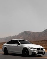 A sleek white sedan is parked on a deserted road with a mountainous landscape in the background. The car has tinted windows and a sporty design, emphasizing its modern appeal. The road stretches into the distance, suggesting a sense of freedom and adventure.
